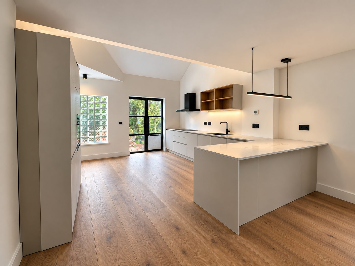 Unit 1 kitchen: pale handleless units and a peninsula on wood floors, with a glass-block window and black-framed glazed doors.